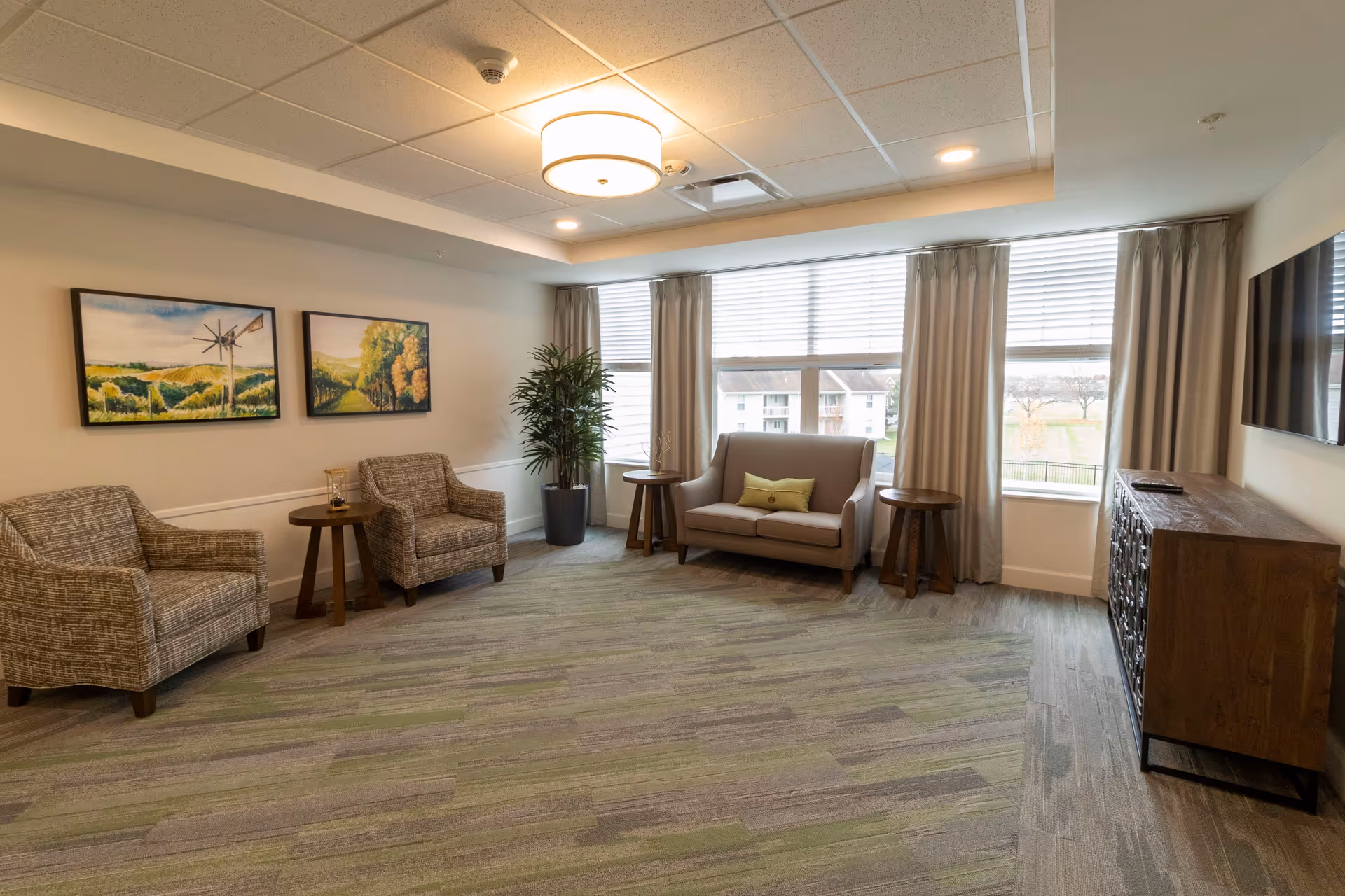 Bright senior living lounge with armchairs, a loveseat, side tables, wall art, and large windows.