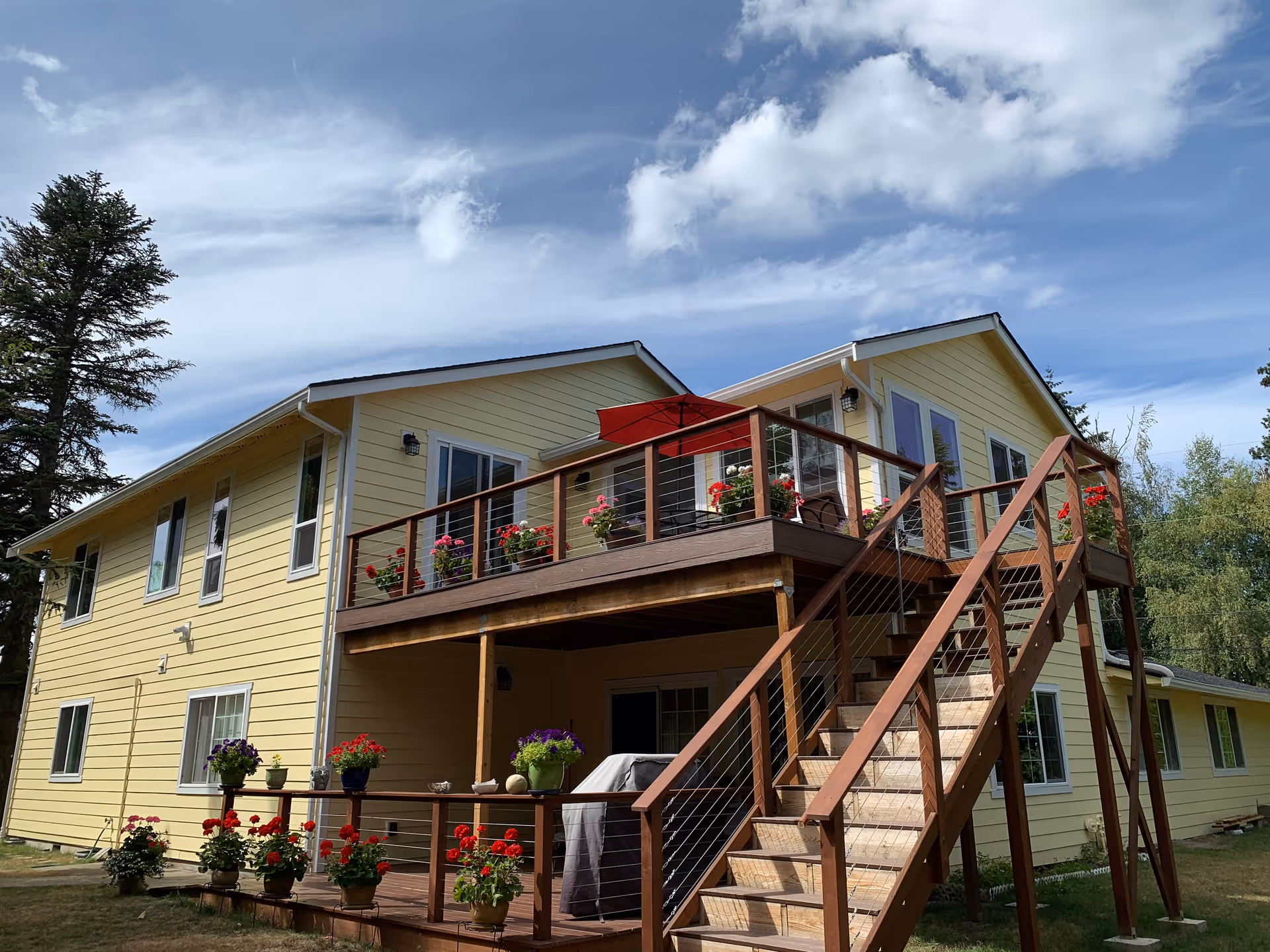 Exterior view of a two-story yellow house with a wooden staircase leading to an upper deck decorated with potted flowers and a red umbrella. The house is surrounded by trees and a grassy yard under a partly cloudy sky.