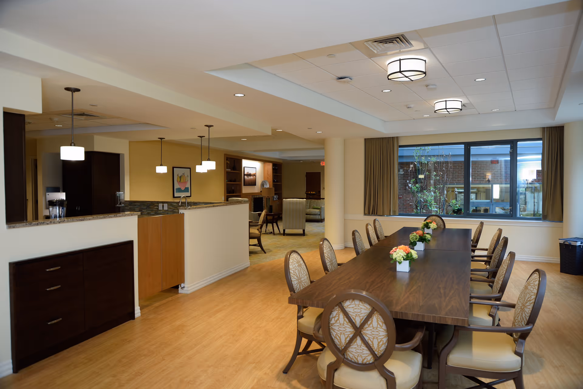 A spacious dining area in a senior living facility with a long wooden table surrounded by upholstered chairs. The table is decorated with small flower arrangements. Large windows with beige curtains allow natural light to enter the room. In the background, there is a kitchen area with pendant lights hanging above the counter and a cozy seating area with armchairs and a fireplace.