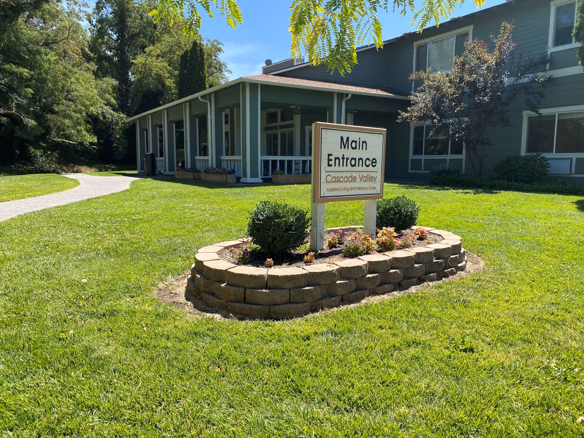 Outdoor view of the main entrance to Cascade Valley Assisted Living and Memory Care facility, featuring a green lawn, a paved walkway, and a sign surrounded by a small flower bed with shrubs and flowers. The building is visible in the background under a clear blue sky.