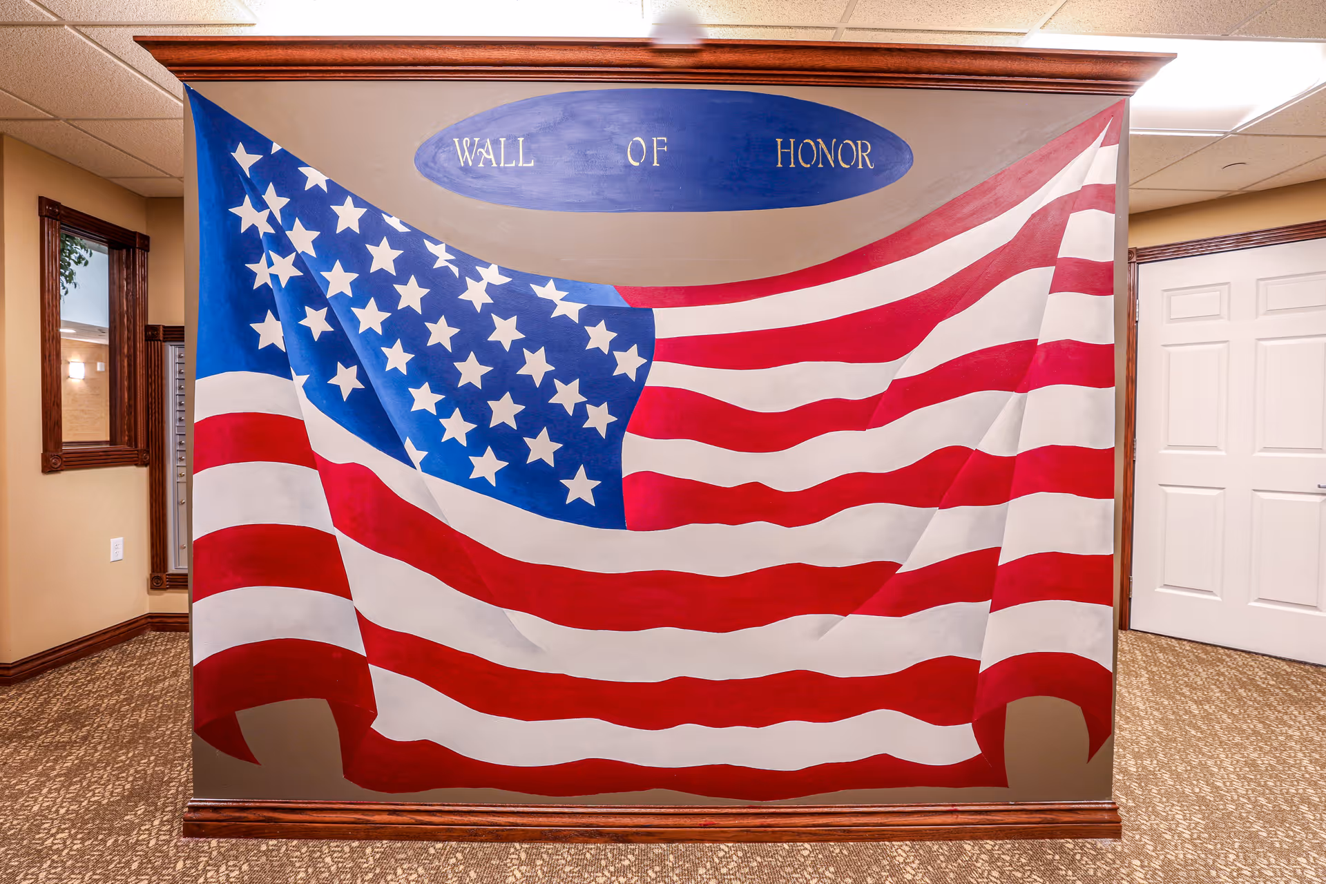 Interior wall mural of a large American flag with the words 'Wall of Honor' painted above it in a senior living facility hallway with beige walls, carpeted floor, and white doors.