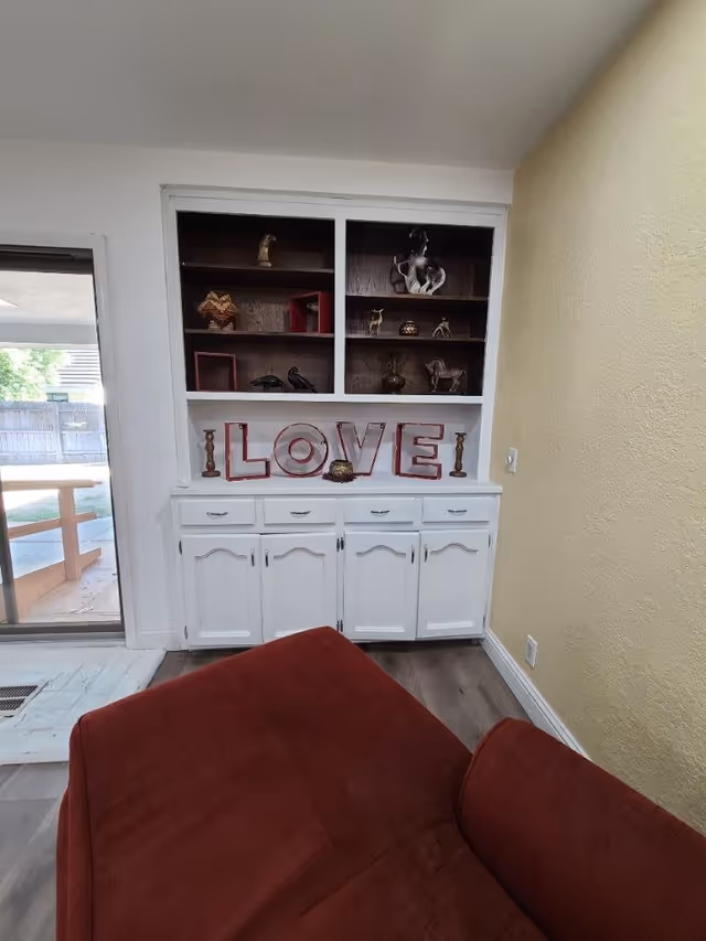 A cozy corner of a living room featuring a built-in white cabinet with shelves displaying decorative items and letters spelling 'LOVE'. A red cushioned chair is partially visible in the foreground. A sliding glass door leads to an outdoor area with a wooden fence and picnic table.