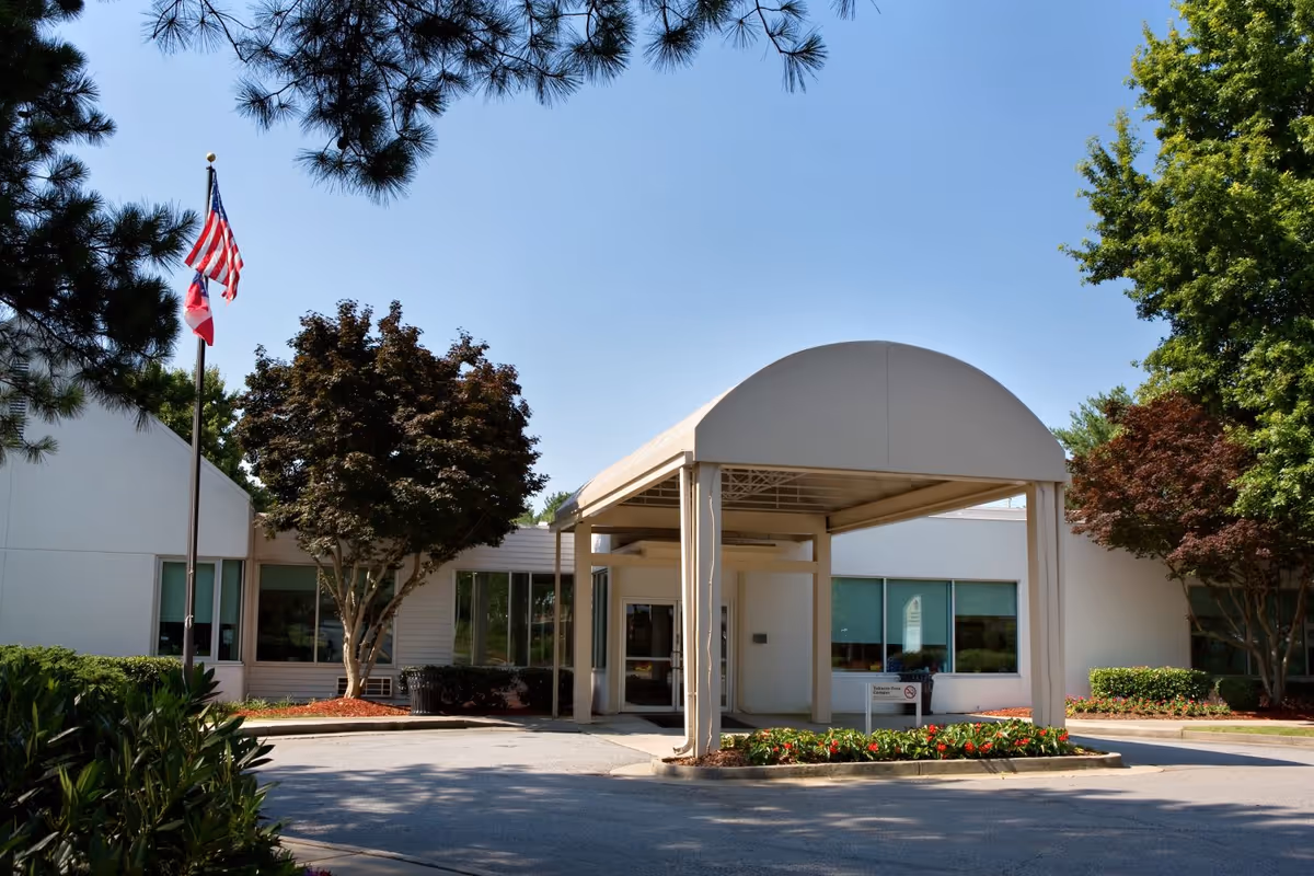 Front exterior view of Northside Gwinnett Extended Care Center showing a covered entrance with a curved roof, surrounded by trees and landscaping under a clear blue sky. Two flagpoles with the American flag and another flag are visible on the left side.