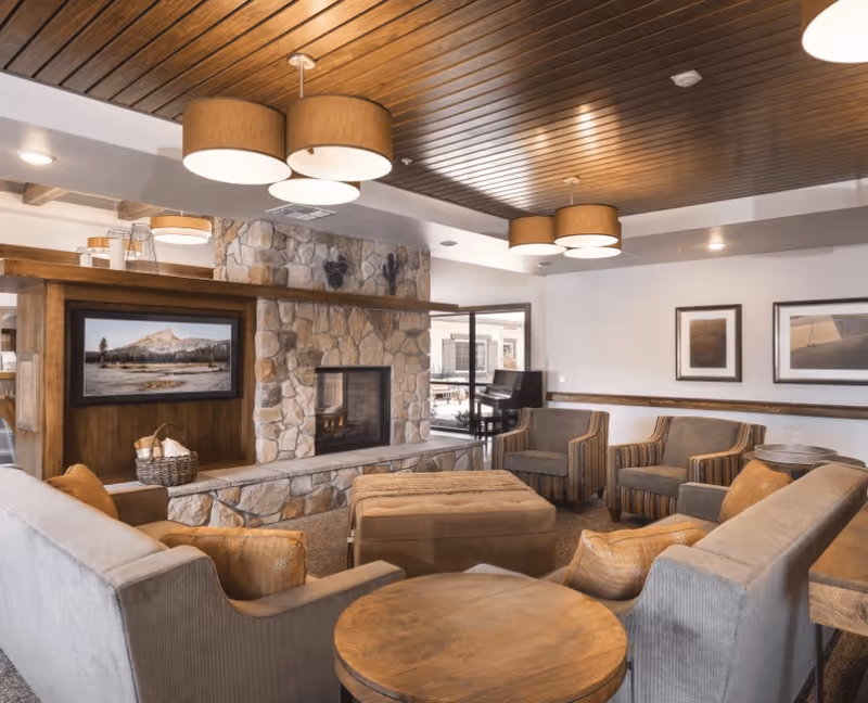 A cozy living room area with a stone fireplace, a mounted TV above the fireplace, and comfortable seating including sofas and armchairs arranged around a large ottoman. The ceiling has wooden paneling with modern drum pendant lights, and there are framed pictures on the walls. A piano is visible in the background near a glass door.