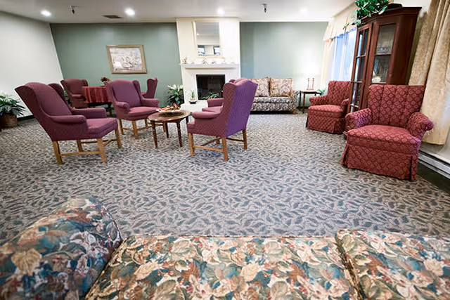 A spacious living room with patterned carpet, several upholstered chairs in purple and red, a floral sofa in the foreground, a wooden coffee table in the center, a fireplace with a mirror above it, and a wooden cabinet near a window with curtains.
