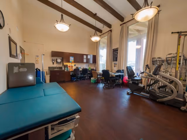 A spacious room with high ceilings and exposed wooden beams, featuring a teal examination table on the left, a desk area with chairs and cabinets along the back wall, large windows with curtains letting in natural light, and exercise equipment including a recumbent bike on the right side.
