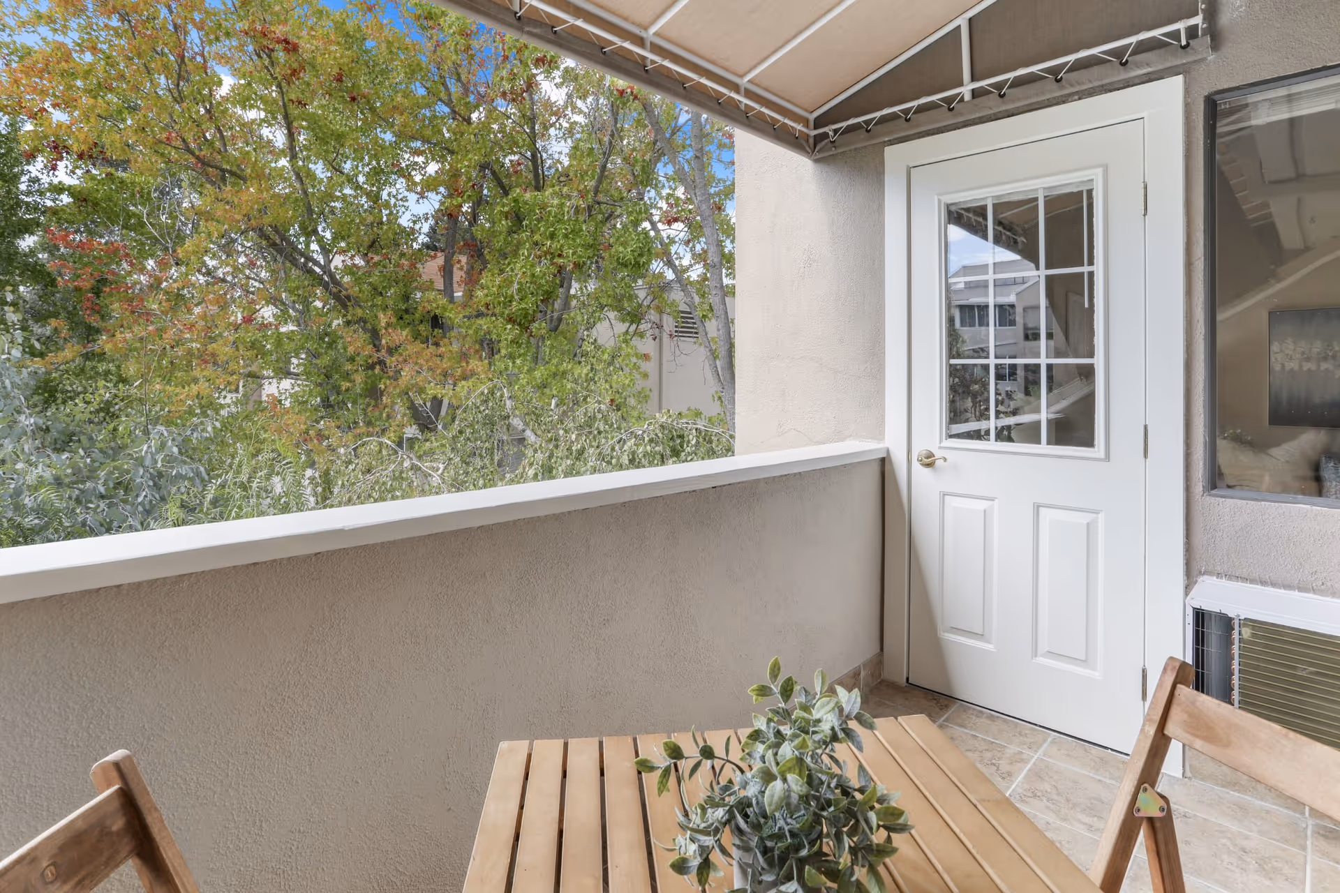 A small outdoor balcony area with a wooden table and chairs. A potted plant is placed on the table. The balcony has a beige stucco wall and railing, with a white door featuring a window with grid panes. Trees with green and some autumn-colored leaves are visible beyond the balcony.