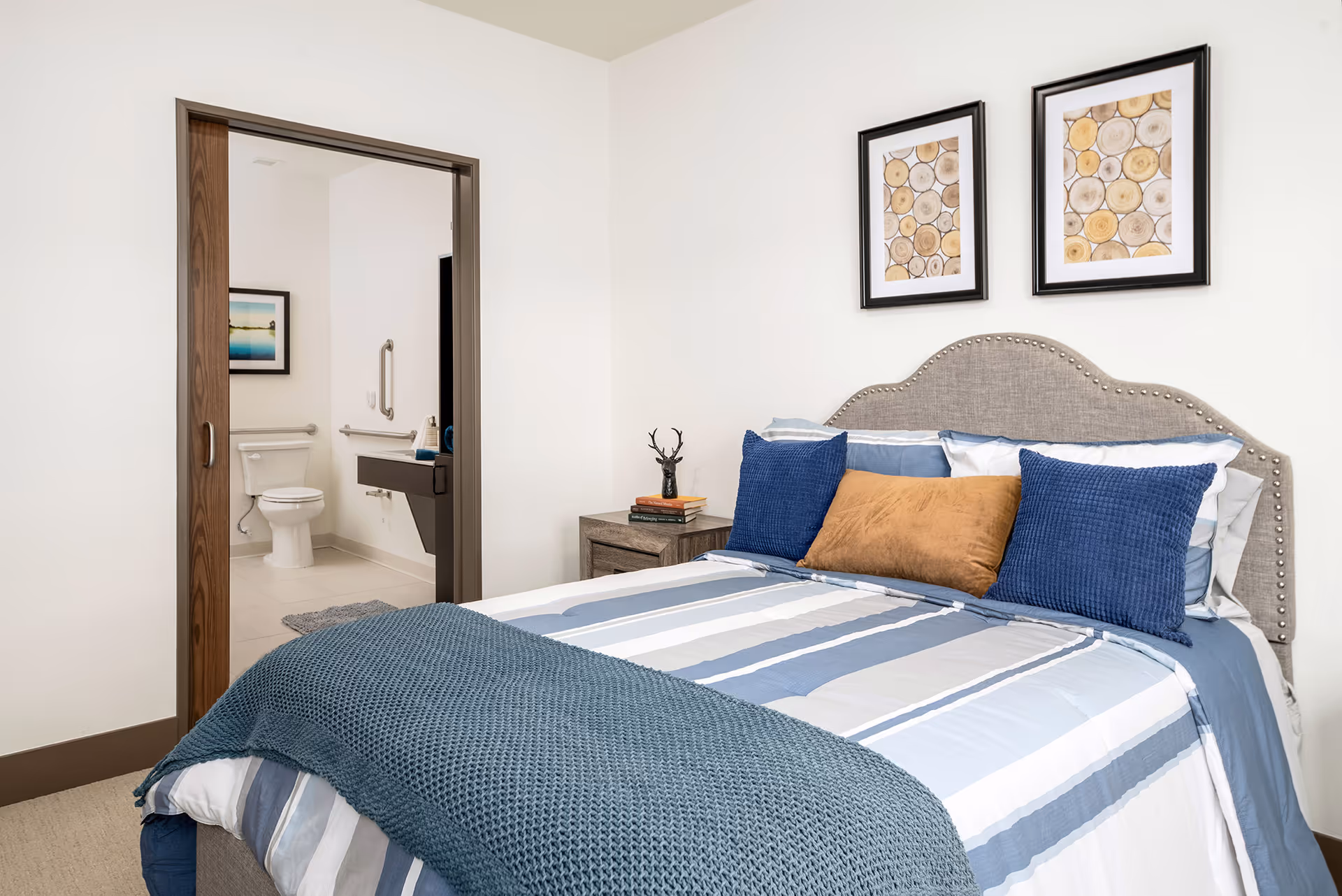 A neatly made bed with blue, white, and gray striped bedding and three pillows, two blue and one brown, in a senior living bedroom. The bed has a gray upholstered headboard with nailhead trim. On the wall above the bed are two framed pictures featuring wood log cross-sections. To the left, there is an open doorway leading to a bathroom with a toilet, grab bars, and a sink visible.
