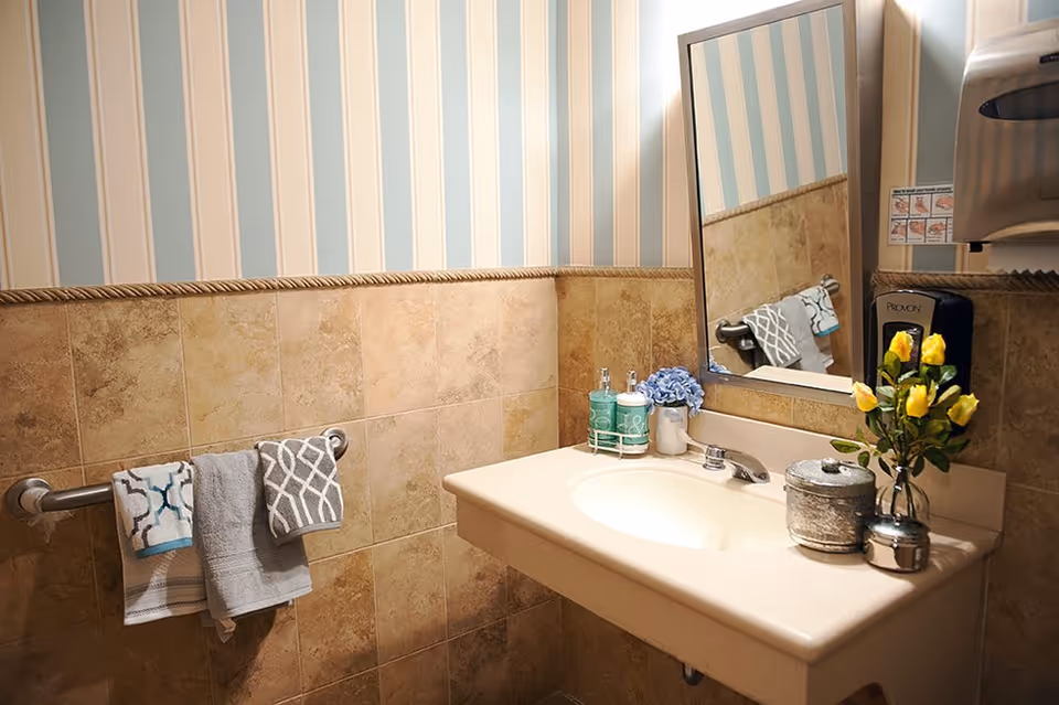 Public bathroom sink area with a mirror, striped wallpaper, tiled walls, hand towels, soap dispensers and a vase of yellow flowers.