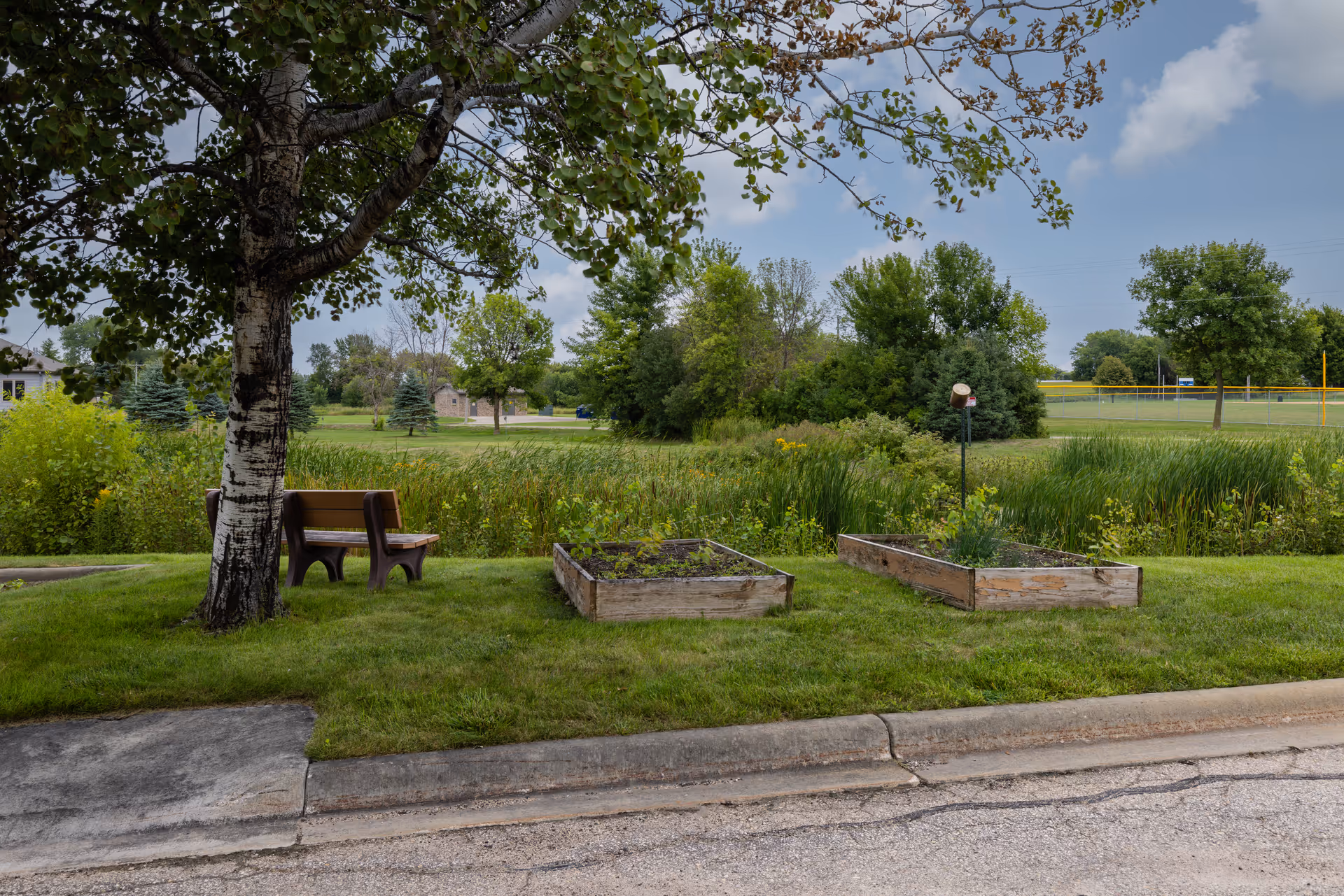 Outdoor garden area with two raised wooden garden beds, a wooden bench under a tree, and a grassy lawn with trees and shrubs in the background under a partly cloudy sky.