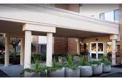 Entrance area of a senior living facility with a covered drop-off zone supported by columns, large potted plants in front, and glass doors leading inside the building.