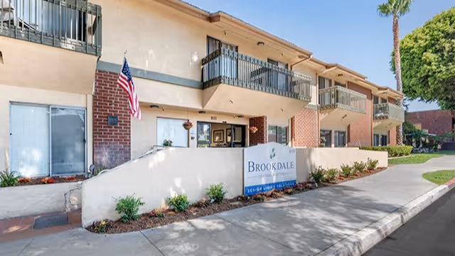 Front exterior of Brookdale Central Whittier showing the entrance sign, balconies, an American flag, and a sidewalk.