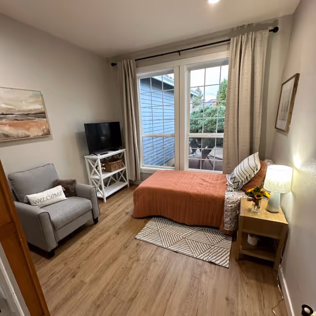 Cozy single bedroom with a daybed covered in an orange throw, an armchair, TV on a stand, nightstand with a lamp, and large windows/door to the outside.