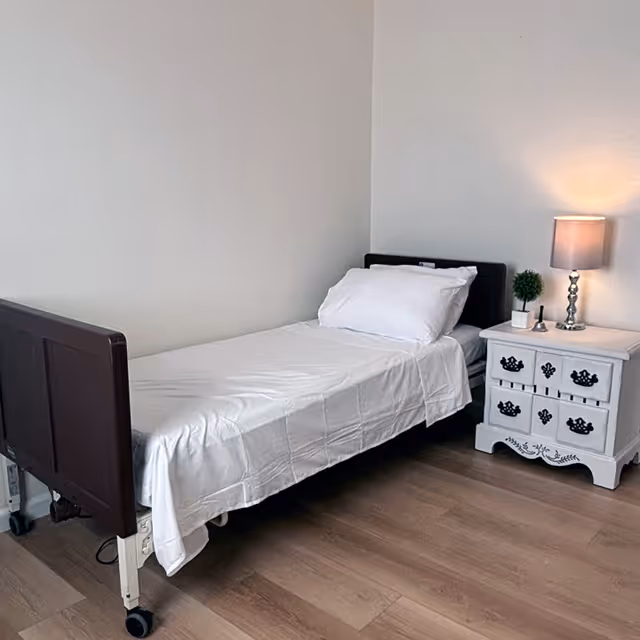 A simple bedroom with a single bed covered in white sheets and a white pillow. Next to the bed is a white nightstand with decorative black handles, a small potted plant, and a lit table lamp. The room has light-colored wooden flooring and plain white walls.