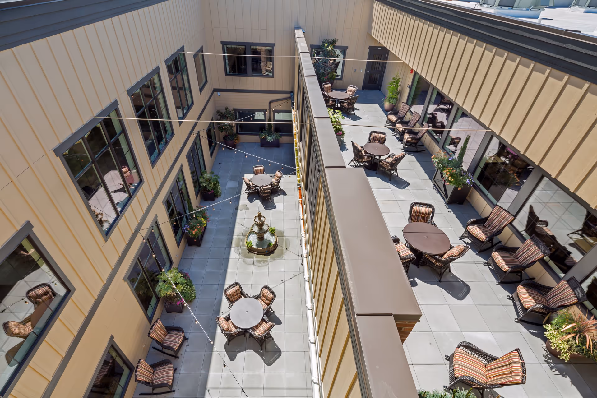Aerial view of an outdoor courtyard area in a senior living facility with multiple round tables and cushioned chairs arranged on a tiled floor. The courtyard is surrounded by beige building walls with large windows and decorated with potted plants and a central water fountain. String lights are hung across the courtyard.