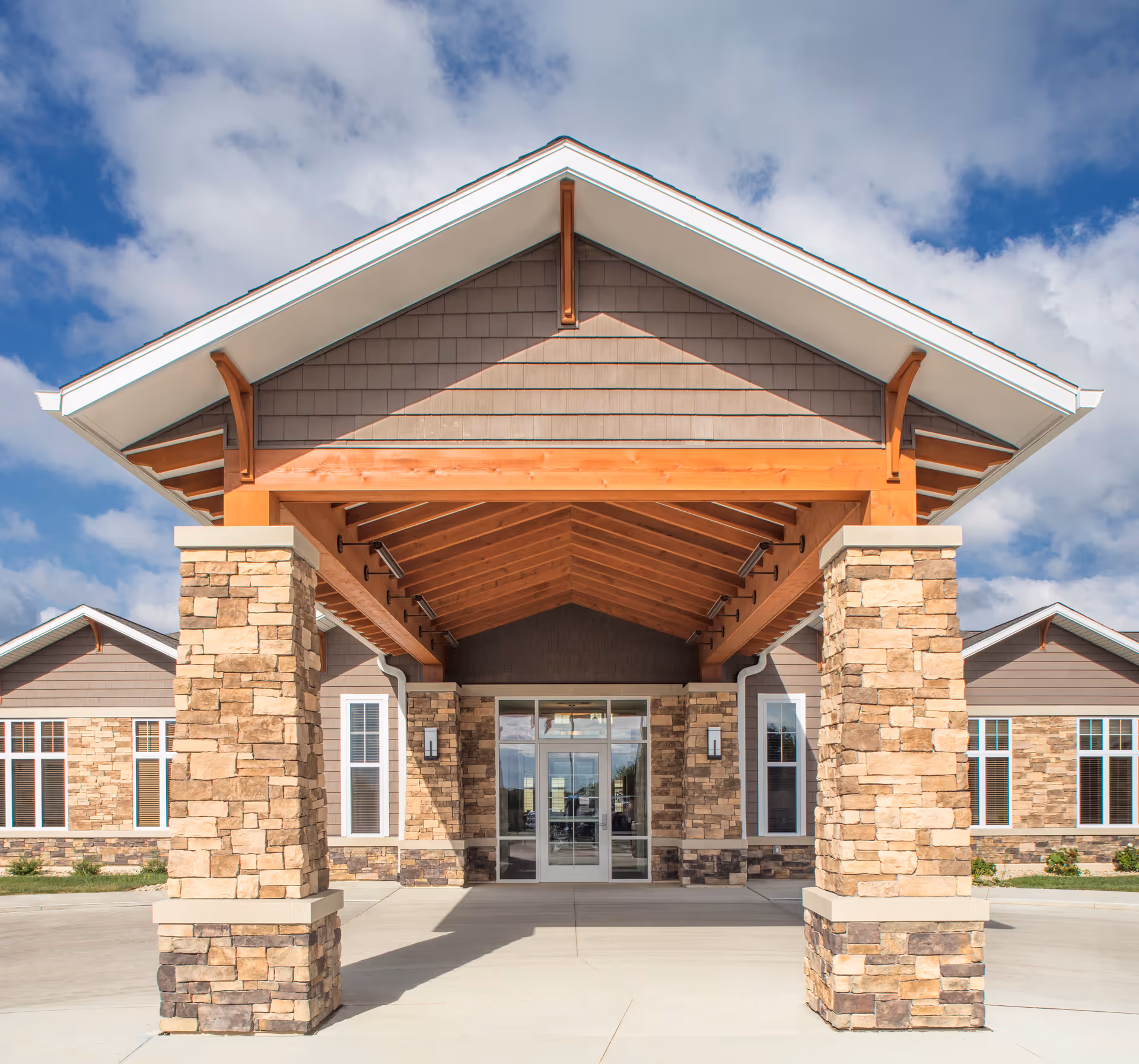Front exterior view of a senior living facility with a covered entrance supported by stone pillars and wooden beams under a partly cloudy sky.