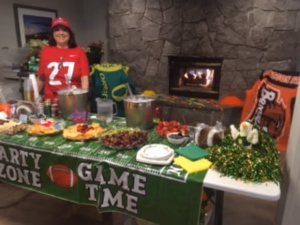 A woman wearing a red sports jersey and cap stands behind a table decorated with a green 'Party Zone Game Time' tablecloth. The table is set with various snacks, drinks, and party decorations. Behind her is a stone wall with a lit fireplace and sports-themed blankets draped over chairs.