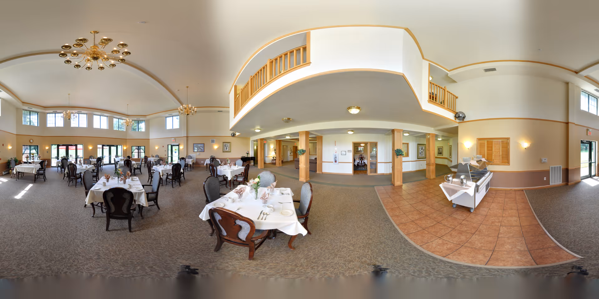 Spacious dining room in a senior living facility with multiple tables covered in white tablecloths, set with plates, glasses, and silverware. The room features high ceilings with chandeliers, large windows allowing natural light, and a serving area with a counter on the right side. The decor includes wooden accents and framed artwork on the walls.