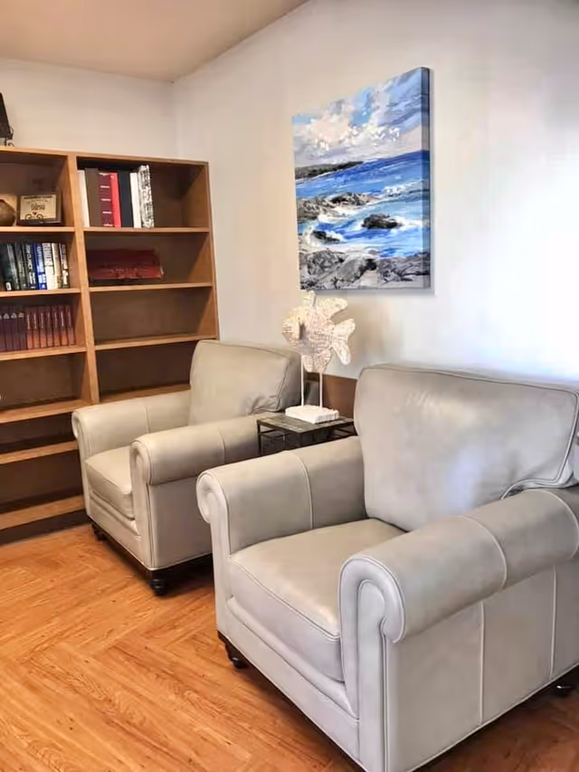 A cozy sitting area with two beige leather armchairs placed side by side on a wooden floor. Behind the chairs is a wooden bookshelf with several books and decorative items. A small side table between the chairs holds a white coral sculpture. Above the chairs, a painting of a rocky shoreline with blue ocean waves and a partly cloudy sky is hung on the wall.