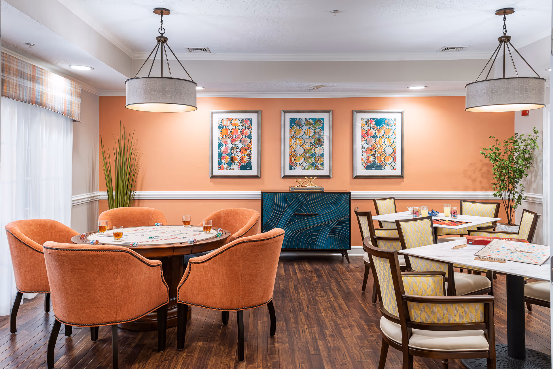 A cozy game room with two tables set up for board games. One round table is surrounded by six orange upholstered chairs with drinks and poker chips on the table. Two square tables with yellow patterned chairs have board games like Scrabble and checkers set up. The room has warm peach-colored walls, three colorful framed artworks, wooden flooring, two hanging light fixtures, and plants in the corners.