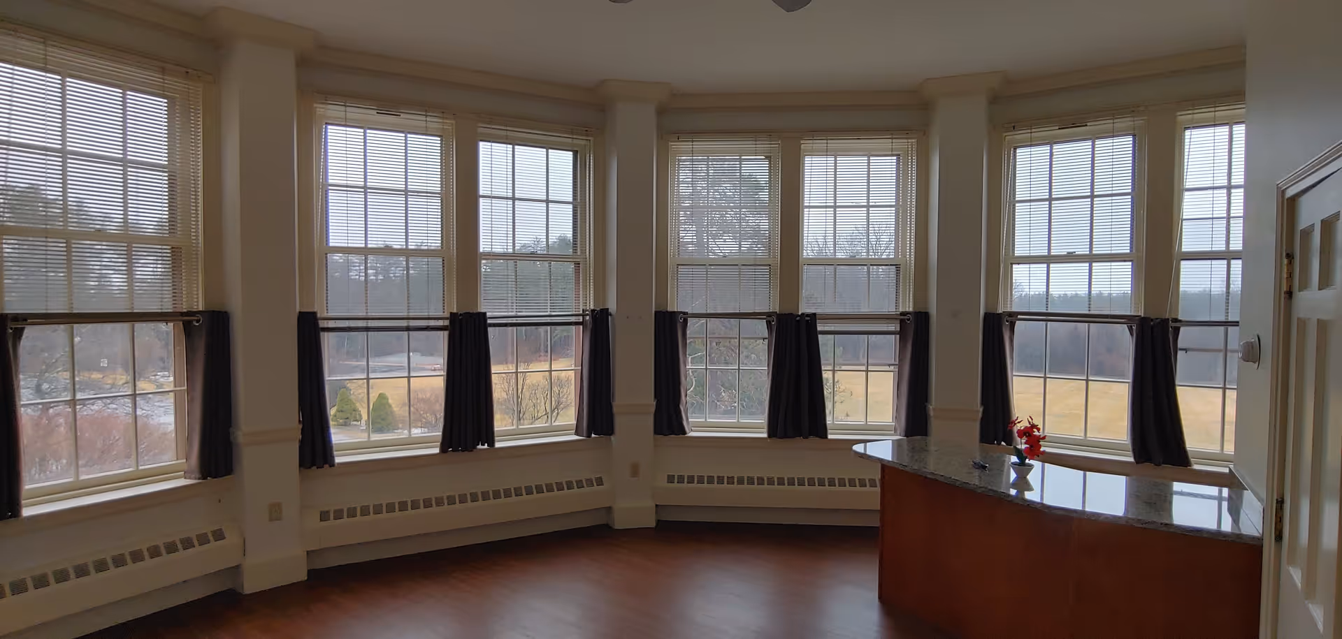 A bright room with large windows featuring white frames and dark curtains halfway drawn. The room has wooden flooring and a curved wooden reception desk with a granite countertop, decorated with a small vase of red flowers. Outside the windows, a grassy landscape and trees are visible.