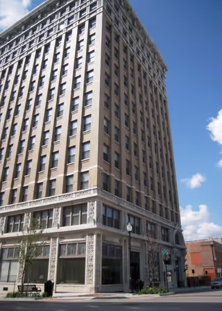 Tall multi-story brick building with decorative stonework on the lower floors, located on a street corner under a blue sky with some clouds.