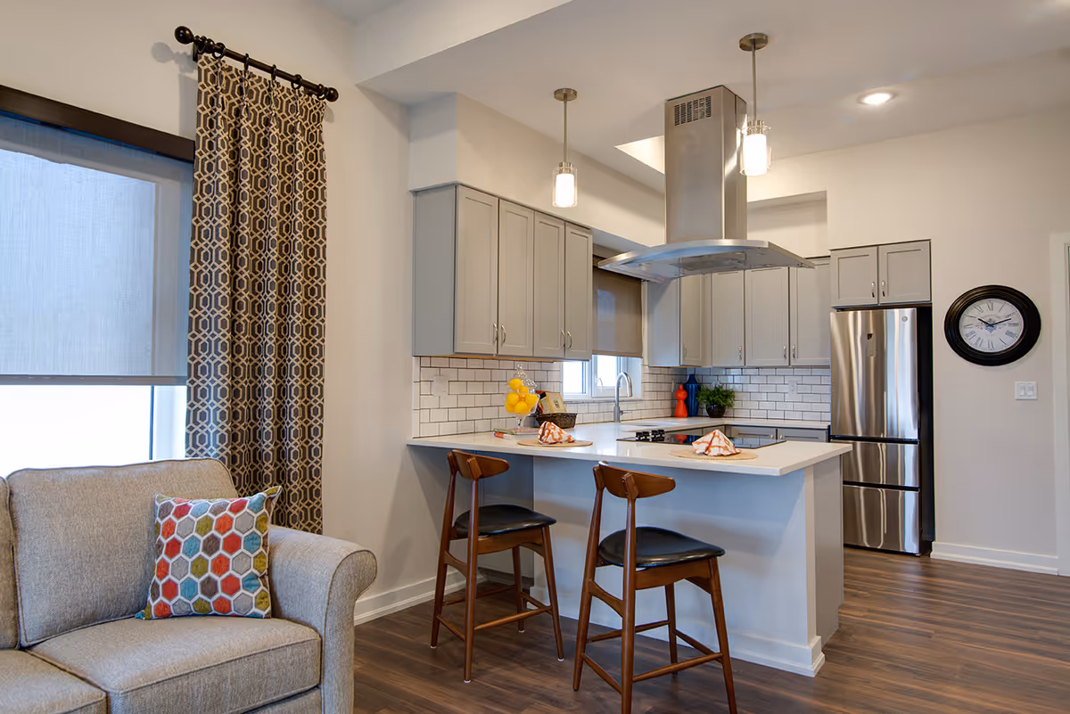 Modern kitchen area with gray cabinets, a white countertop island with two wooden bar stools, stainless steel refrigerator, and a range hood. Adjacent to the kitchen is a beige armchair with a colorful hexagonal patterned pillow and patterned curtains on the window.