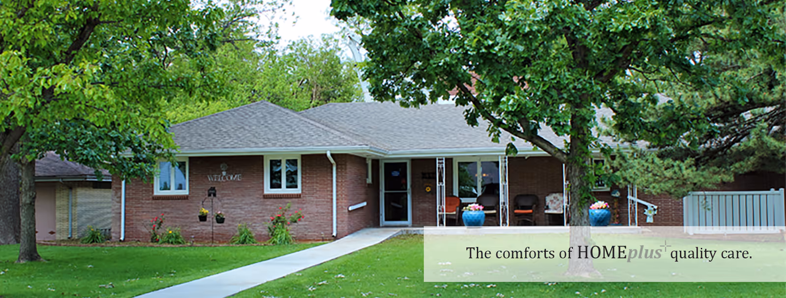 Single-story brick building with a gray roof, surrounded by green grass and trees. A concrete walkway leads to the entrance, which has glass doors and a small covered porch with chairs and potted plants. The word 'WELCOME' is visible on the building's wall near the entrance.