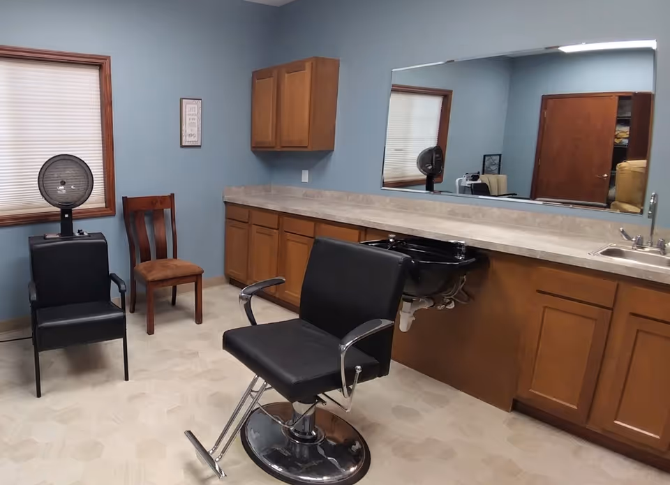Interior of a salon room with a black salon chair in front of a counter with a sink and a black wash basin. There is a large mirror above the counter, a wooden chair, a black chair with armrests, and a hair dryer near a window with blinds. The walls are painted blue and there are wooden cabinets below and above the counter.