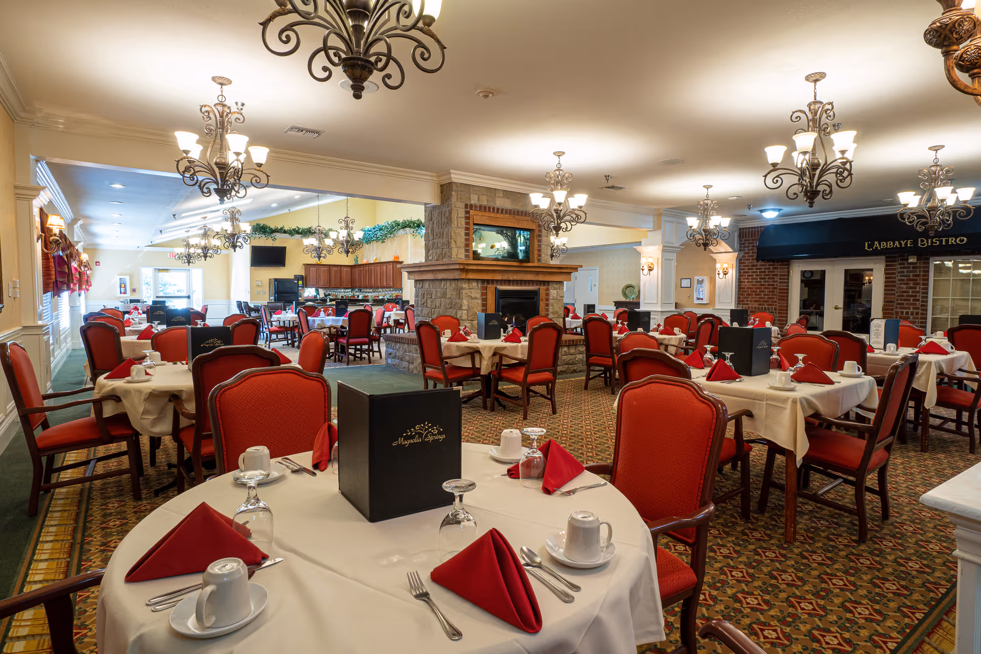 Well-appointed dining room with multiple round tables covered in white linens, red napkins and chairs, chandeliers, and a central stone fireplace.