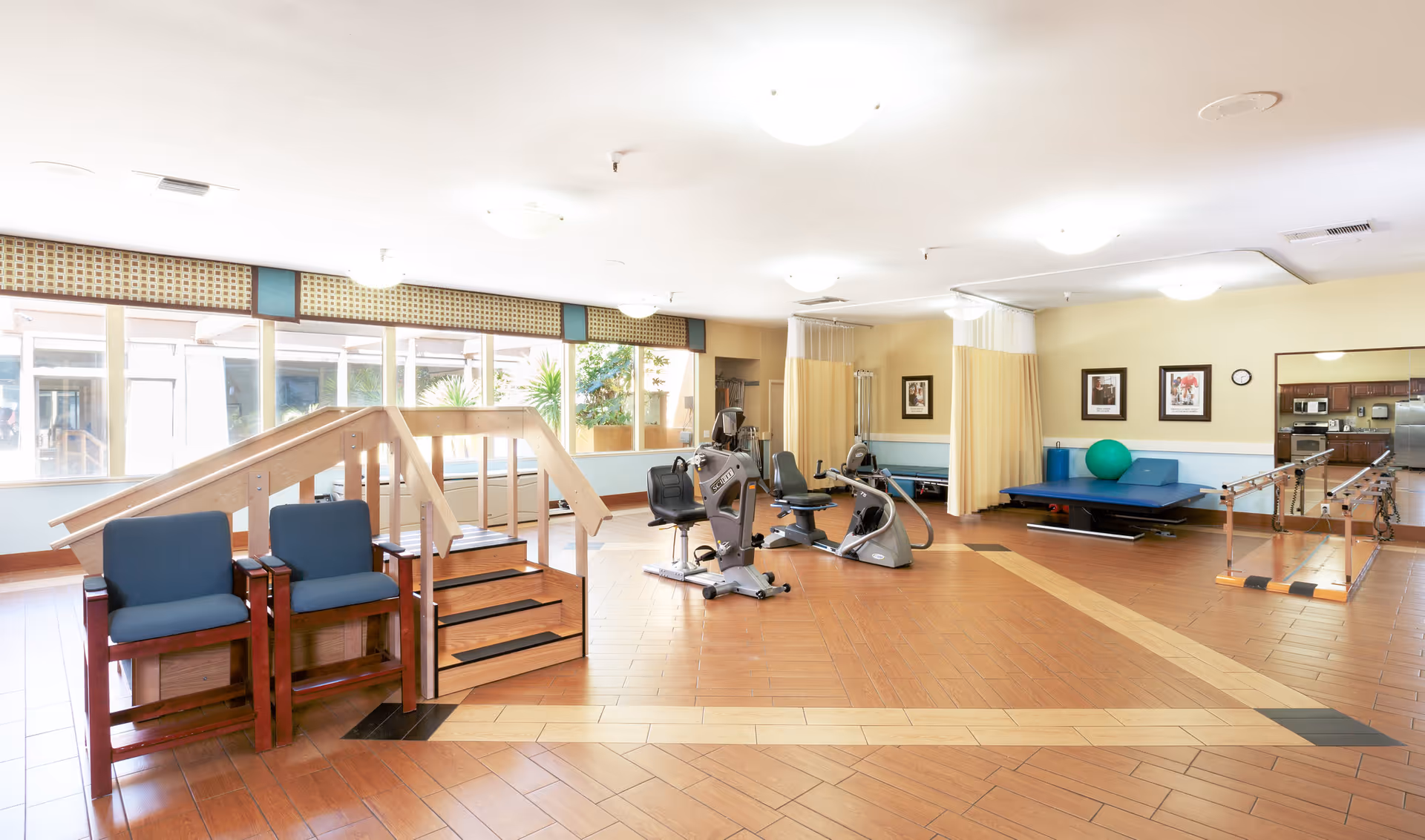 A spacious rehabilitation or physical therapy room with exercise equipment including stationary bikes, parallel bars for walking practice, a small wooden staircase with handrails, two blue chairs, and a padded therapy table with a large green exercise ball. The room has large windows letting in natural light and light-colored walls with framed pictures.