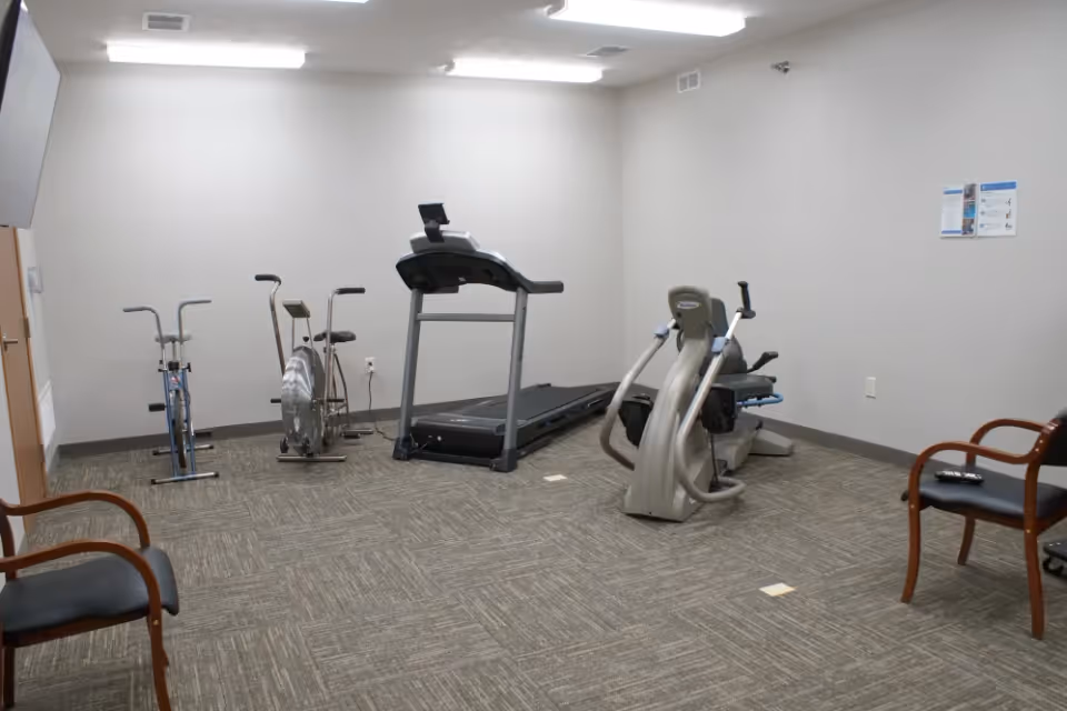 Small fitness room with a treadmill, stationary bikes, a recumbent exercise machine and chairs on a carpeted floor.