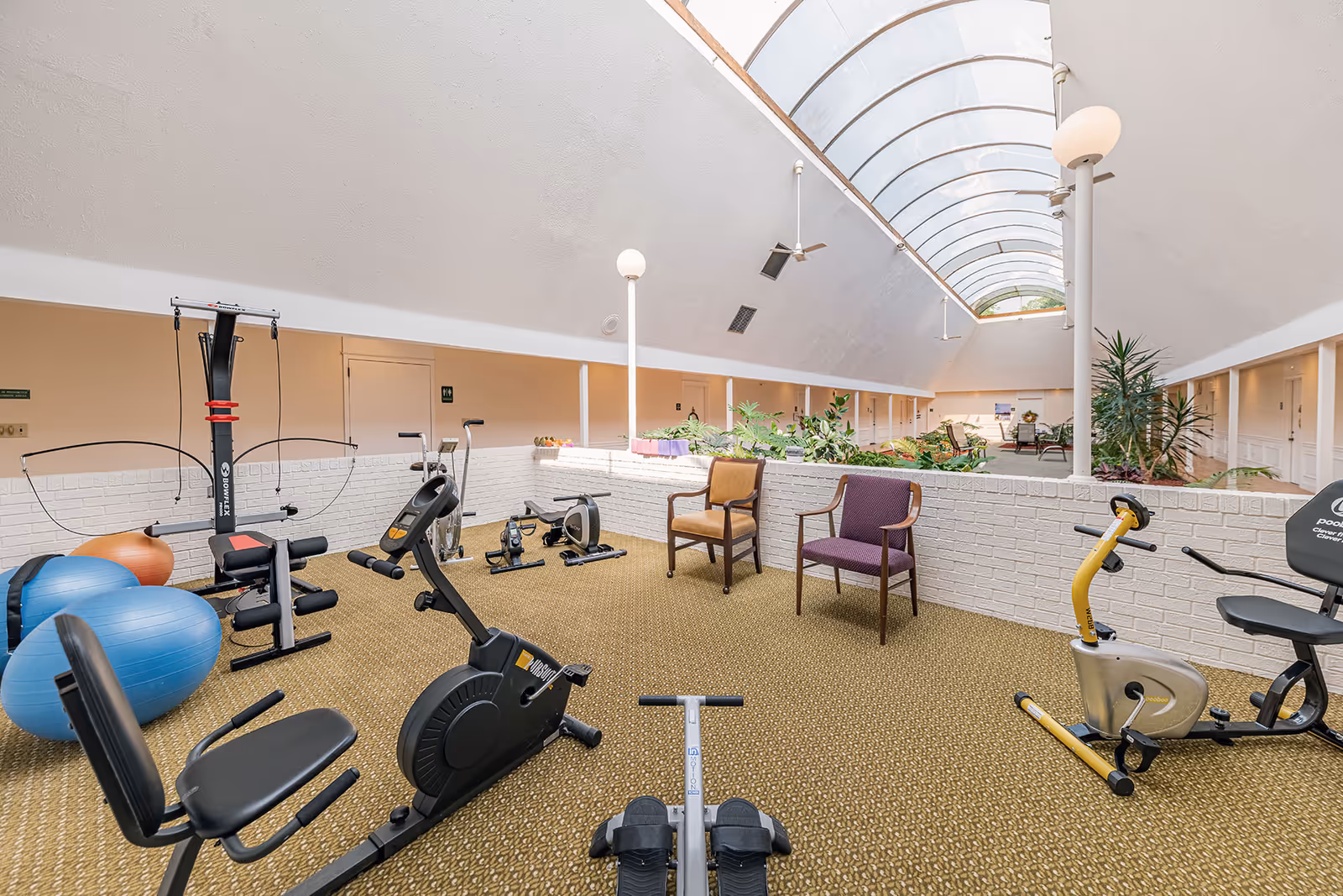 Indoor exercise area in a senior living facility with various fitness equipment including stationary bikes, a rowing machine, exercise balls, and a multi-gym machine. The space has a high arched skylight ceiling allowing natural light, beige carpeted floor, white brick half walls, and two chairs placed near the equipment. There are plants and seating areas visible in the background along a corridor with multiple doors.