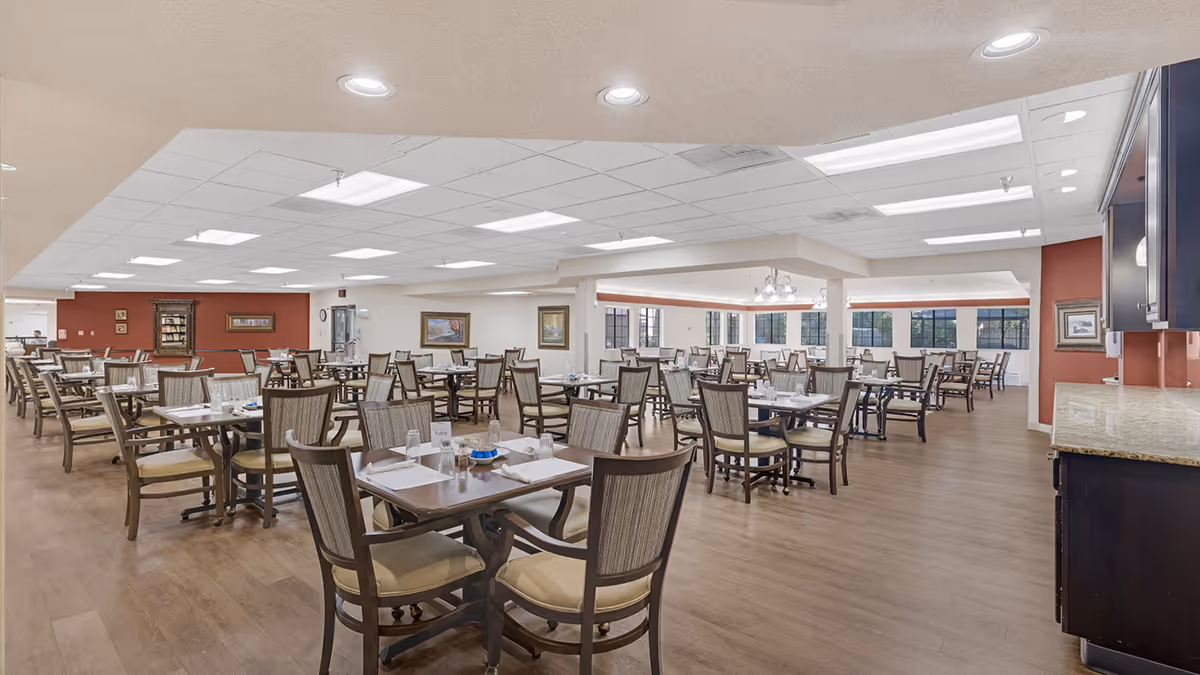 Spacious dining room in The Cambridge Senior Living facility with multiple wooden tables and chairs arranged neatly. The room has large windows allowing natural light, recessed ceiling lights, and framed artwork on the walls. The floor is wooden, and there is a countertop area on the right side.