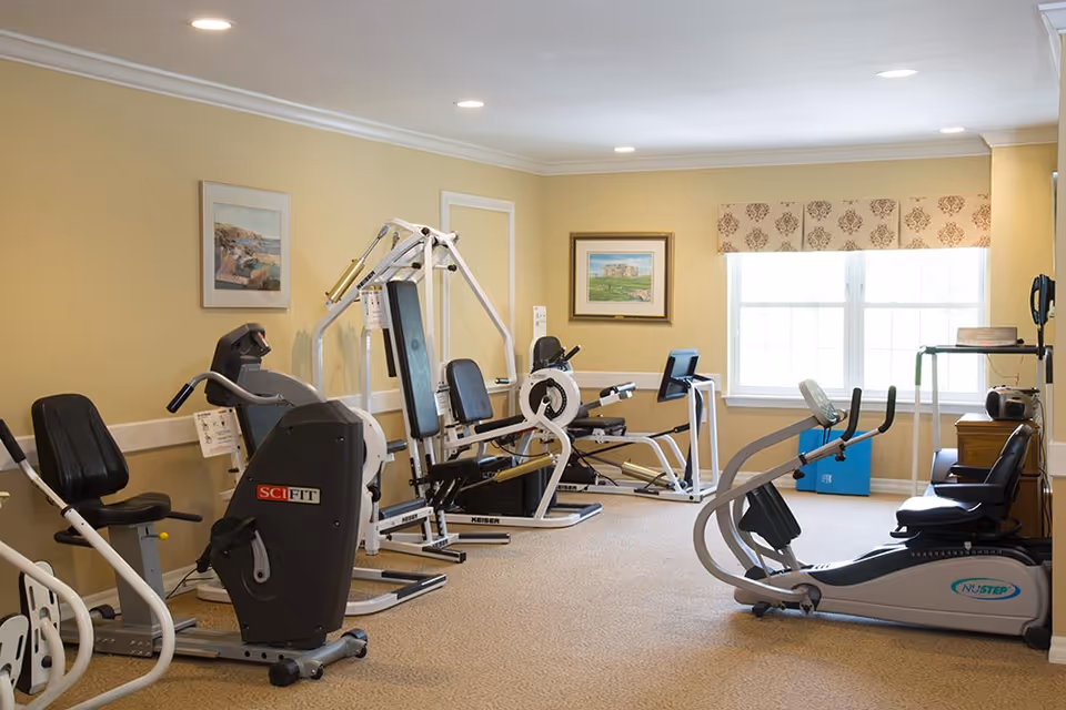A well-lit fitness room in a senior living facility featuring various exercise machines including stationary bikes, a rowing machine, and a multi-function strength training machine. The room has beige walls with framed artwork, a large window with patterned valance, and carpeted flooring.