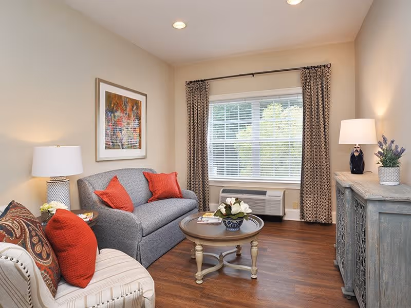 Cozy living room with a gray sofa and armchair, round coffee table, lamps, and a window with blinds and curtains.