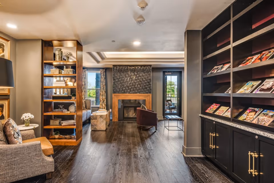 A cozy living room area in a senior living facility featuring a fireplace with a decorative tile surround, a bookshelf filled with magazines on the right, a wooden shelving unit with decorative items on the left, comfortable seating including a gray armchair and a purple chair, and large windows with patterned curtains allowing natural light to enter.