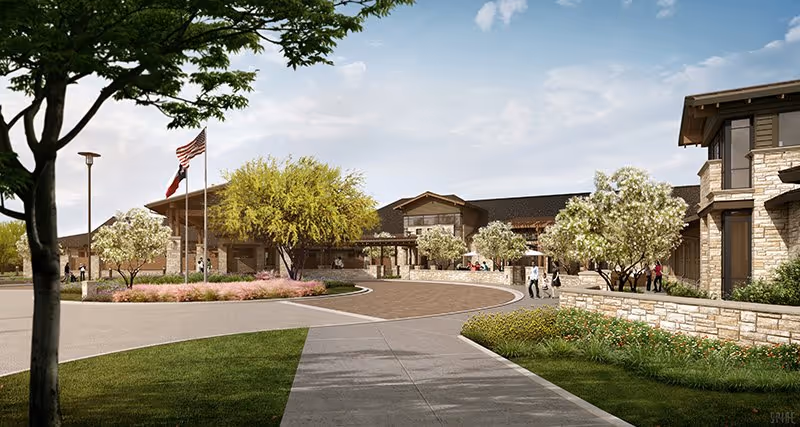 Exterior view of a senior living facility with a circular driveway, landscaped garden areas with trees and flowers, and a building with stone and wood accents under a partly cloudy sky.
