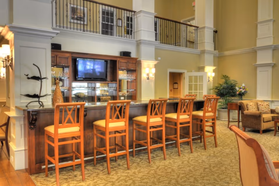 A spacious and well-lit common area with a long wooden bar counter and six high wooden chairs with cushioned seats. Behind the bar is a wooden cabinet with glass shelves holding glassware and a mounted flat-screen TV. The room features high ceilings, beige walls, decorative columns, wall sconces, and a carpeted floor. There is also a seating area with upholstered chairs and a small table with a plant and flowers.