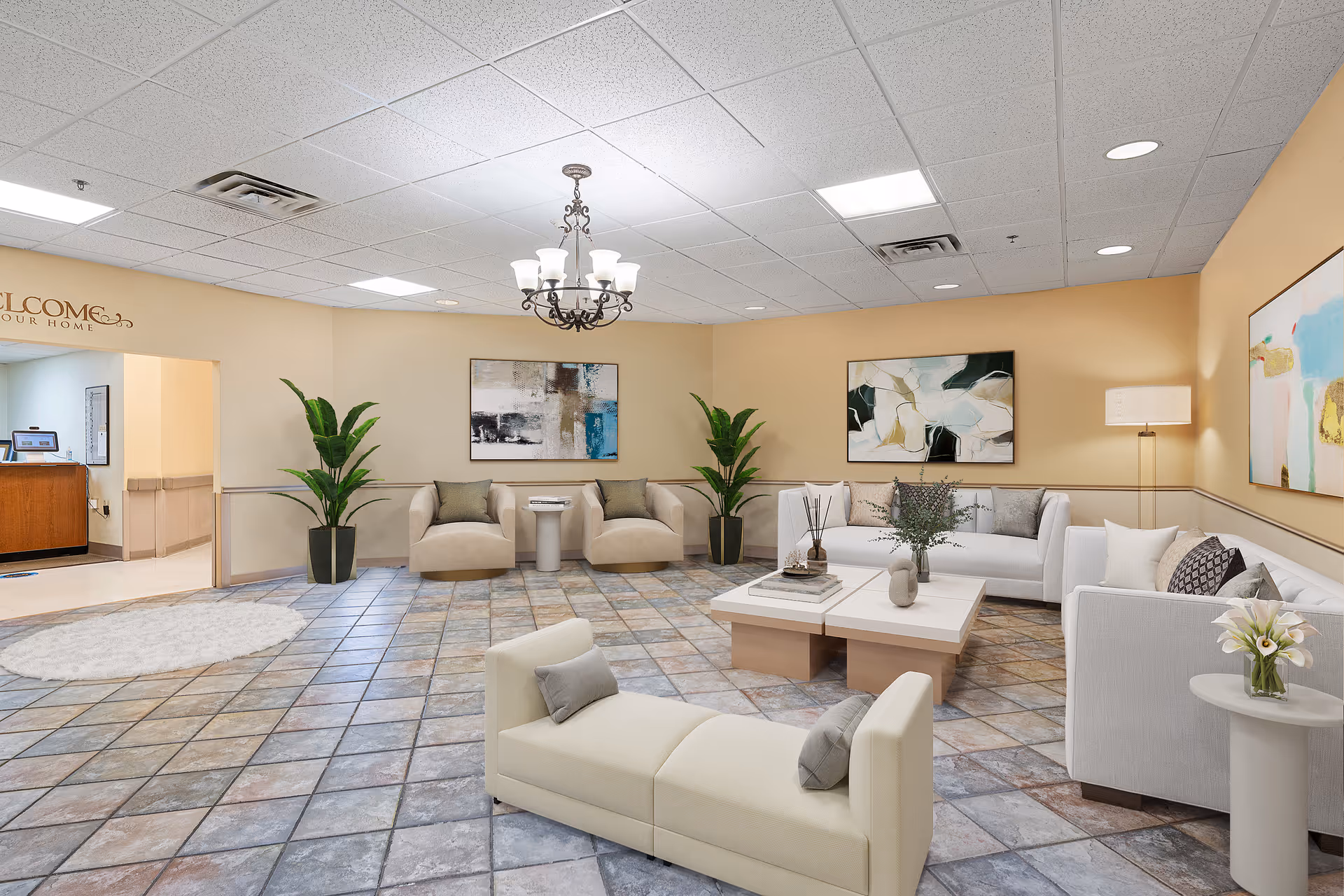A spacious and well-lit living room area with beige walls and tiled flooring. The room features modern furniture including two beige armchairs with a small table between them, a white sectional sofa, a white loveseat, and a white coffee table with decorative items. There are two large potted plants, three abstract paintings on the walls, a chandelier hanging from the ceiling, and a floor lamp in the corner. An open doorway leads to a reception area with a wooden desk and computer.