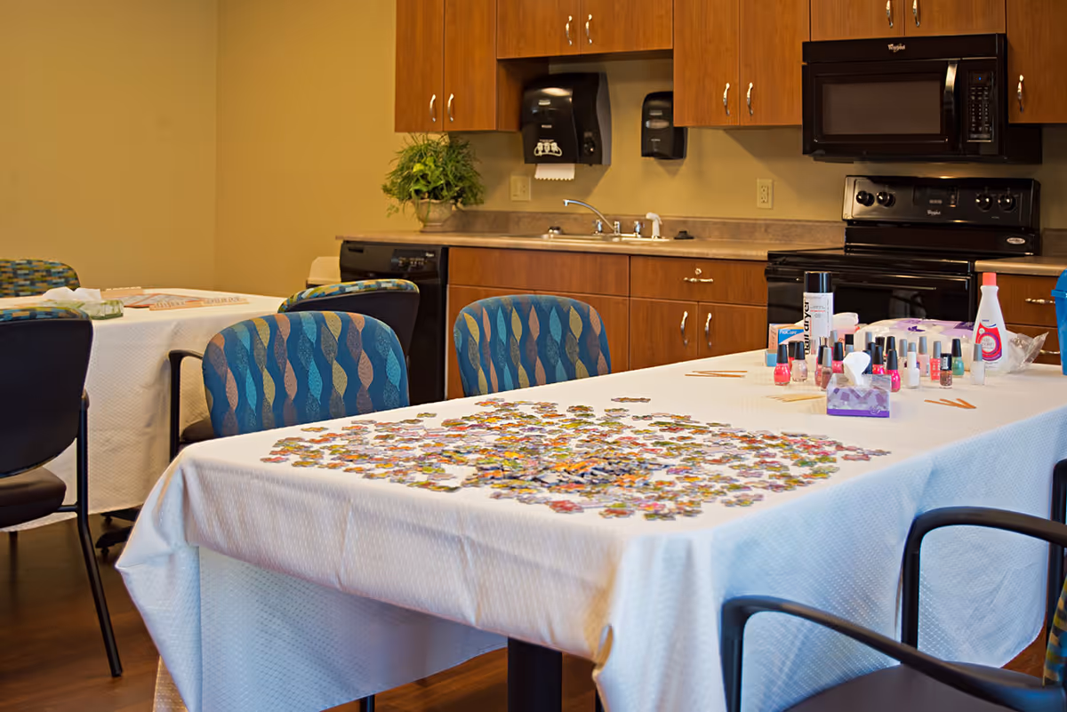 Interior room with tables covered in white tablecloths, one table has a partially completed jigsaw puzzle and various nail polish bottles and manicure supplies. The room has wooden cabinets, a black microwave, stove, dishwasher, and a sink with paper towel and soap dispensers mounted on the wall.