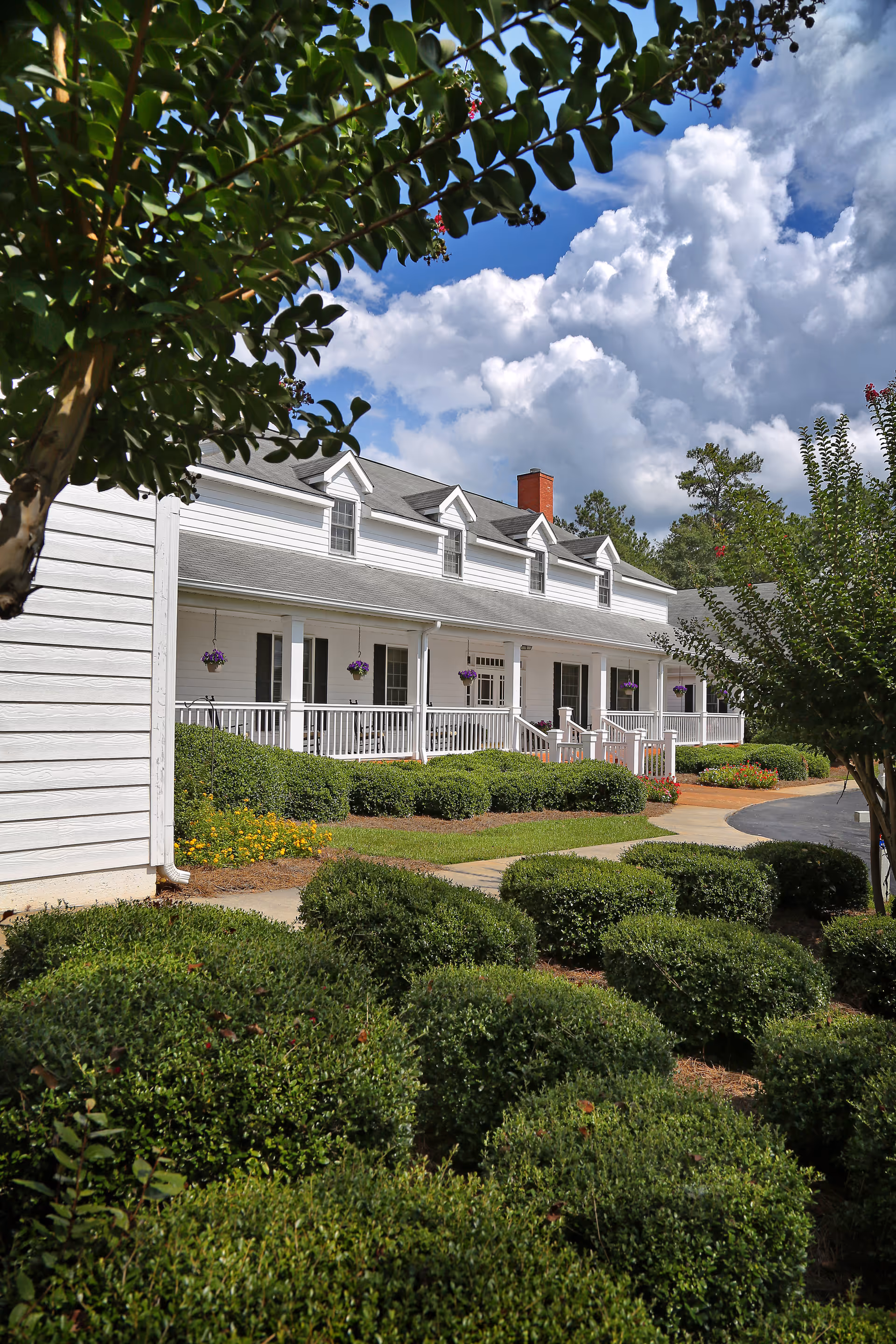Exterior view of a white senior care facility building with a porch, surrounded by neatly trimmed bushes and trees under a partly cloudy blue sky.