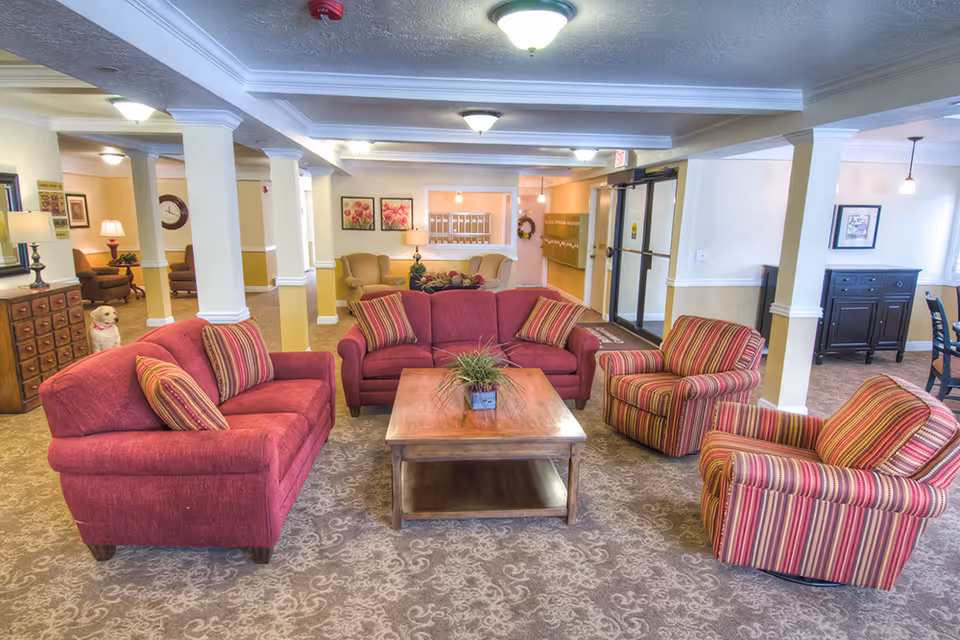 A cozy and well-lit common area in a senior living facility featuring two red sofas and two striped armchairs arranged around a wooden coffee table with a small plant. The room has beige walls with white trim, carpeted floors, and several ceiling lights. In the background, there are additional seating areas, framed floral artwork, a set of mailboxes, and a glass exit door. A small dog is visible near a wooden chest of drawers on the left side.