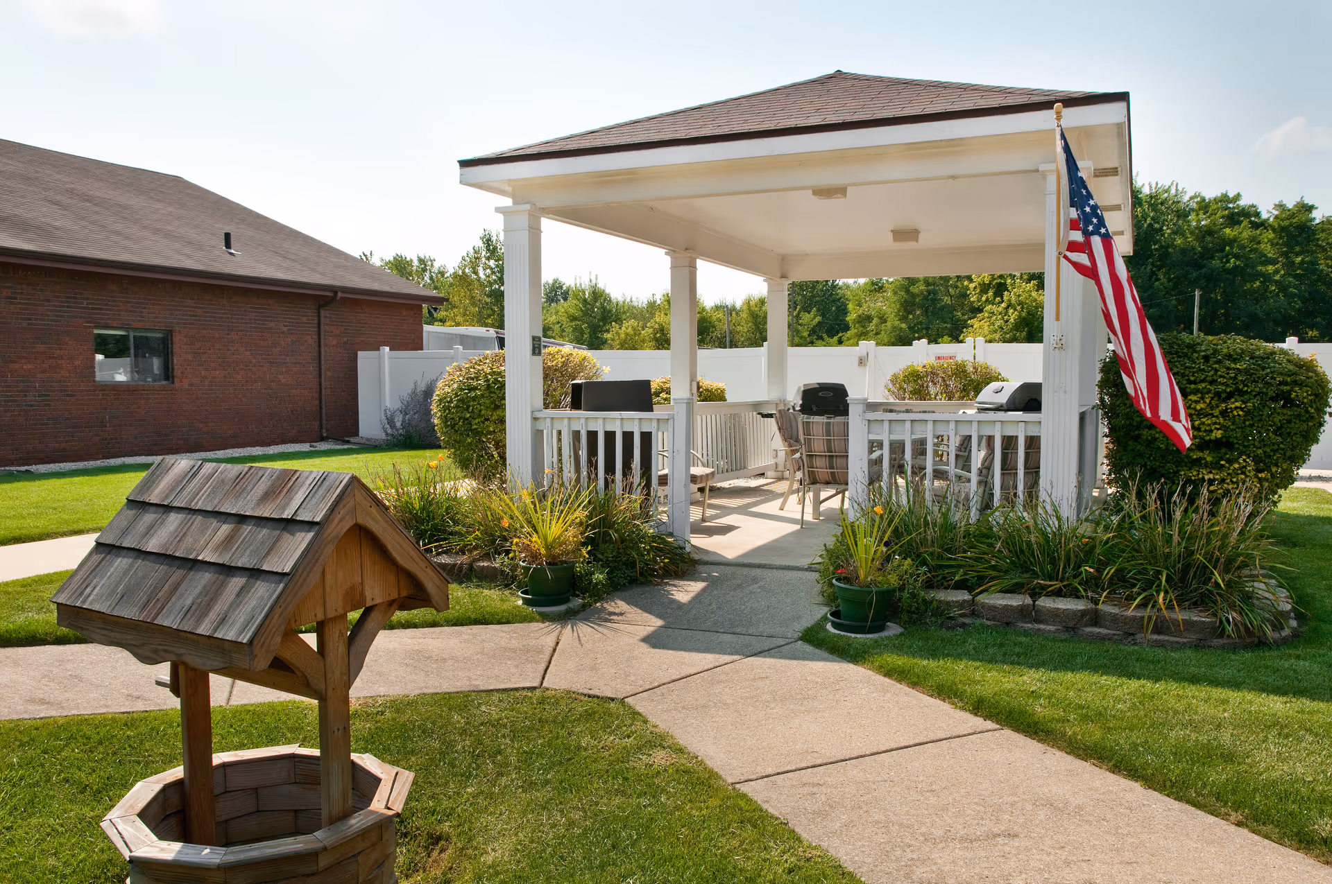 Outdoor gazebo area with white pillars and roof, surrounded by greenery and plants. There are chairs and grills inside the gazebo, and an American flag is mounted on one of the pillars. A wooden wishing well structure is visible in the foreground on a grassy lawn with paved walkways.