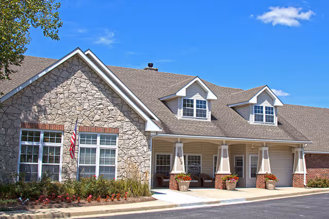 Exterior view of a senior living facility building with stone and siding facade, multiple windows, a covered entrance with columns, potted plants, and an American flag, under a clear blue sky.