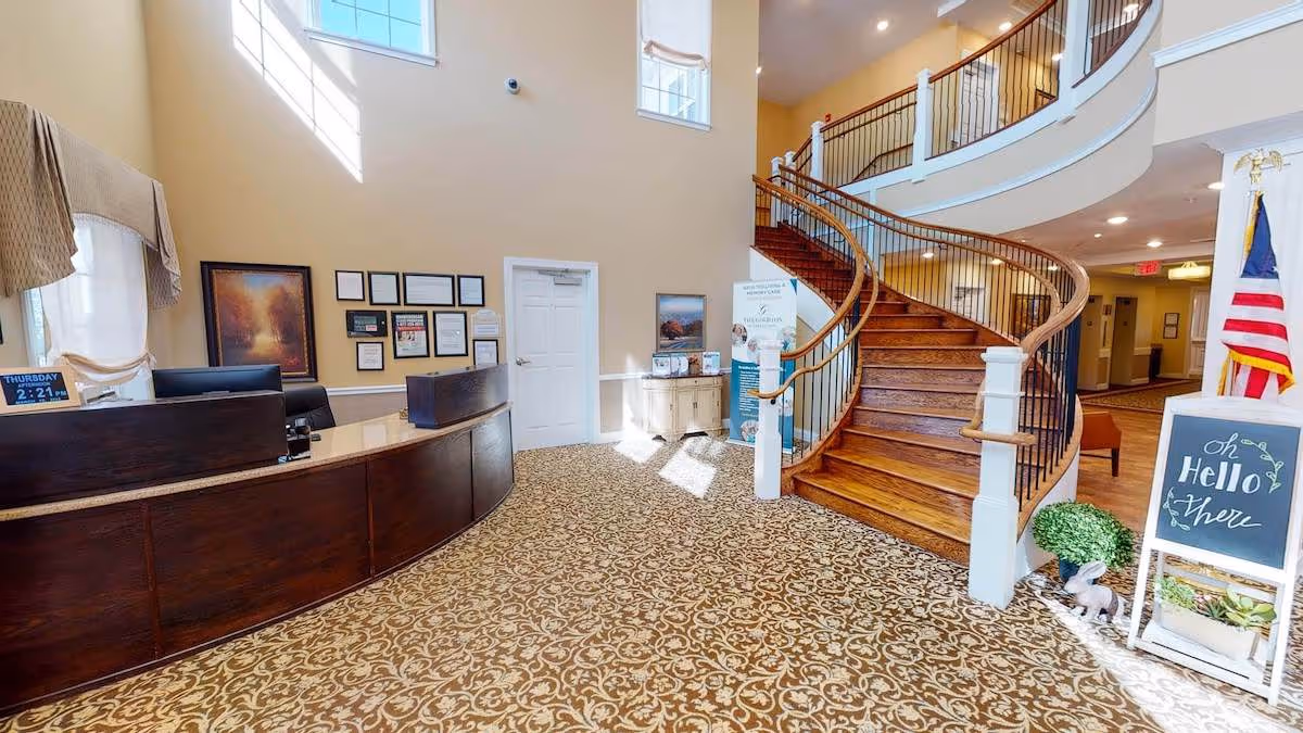 Interior view of a senior living facility lobby with a curved wooden staircase, a dark wood reception desk, patterned carpet, framed pictures on the wall, and a small chalkboard sign that says 'oh Hello there'. There is also an American flag and some decorative plants near the staircase.