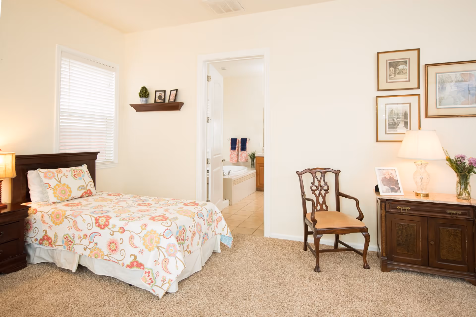 A cozy senior bedroom with a single bed covered in a floral patterned quilt, a wooden nightstand with a lamp, a wooden chair, and a wooden cabinet with a lamp, framed pictures, and a vase of flowers. The room has beige carpet and cream-colored walls. An open door reveals a bathroom with a bathtub, towels, and a sink.