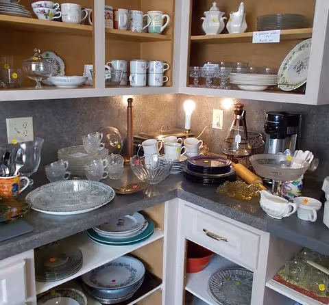 A kitchen corner with open shelves and cabinets filled with various dishes, cups, glassware, and plates. A countertop holds more glassware, a coffee maker, and a lit light bulb. The shelves contain neatly arranged mugs, bowls, and plates.