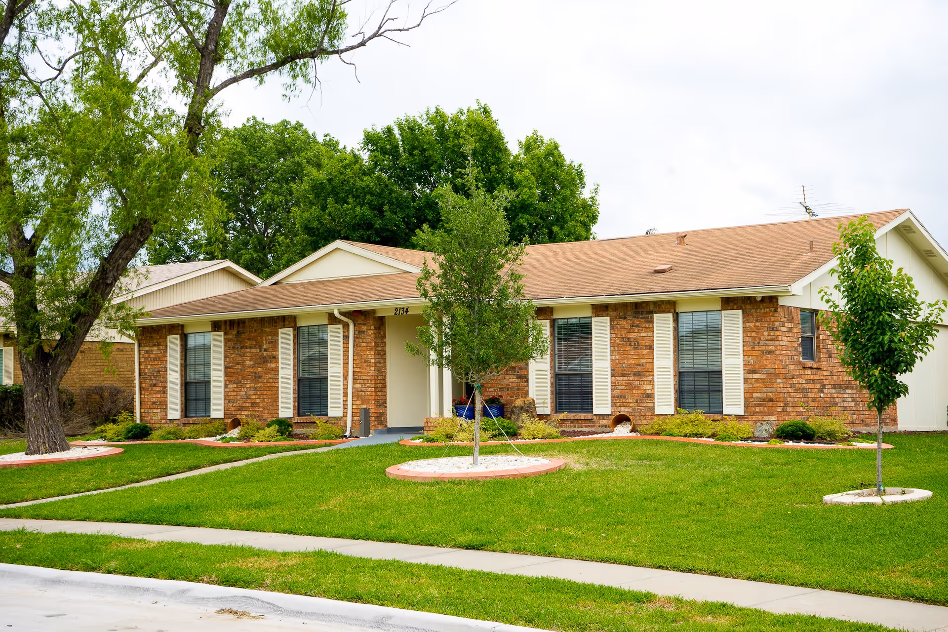 Single-story brick building with a brown roof surrounded by green grass, small trees, and shrubs under a cloudy sky.