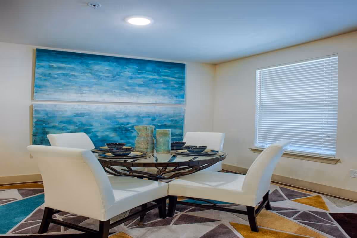 A modern dining area with a round glass table set with four place settings and two decorative vases in the center. The table is surrounded by four white upholstered chairs. The room has a large window with closed blinds and two large blue abstract paintings on the wall. The floor features a colorful geometric patterned rug.